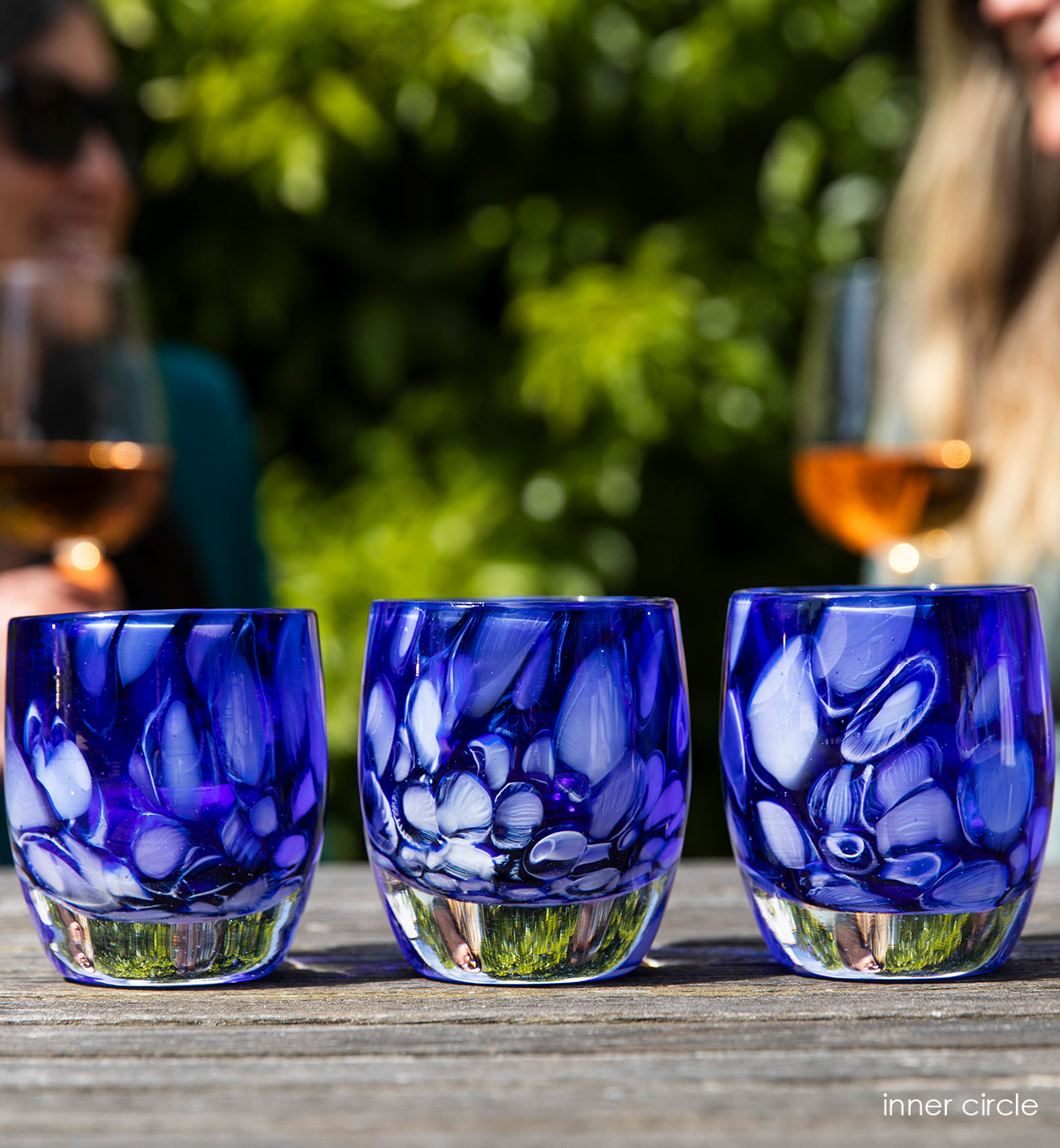 three inner circle, translucent dark blue and white murrini hand-blown glass votive candle holders on a wood outdoor table, with two friends smiling and enjoying wine in the background.