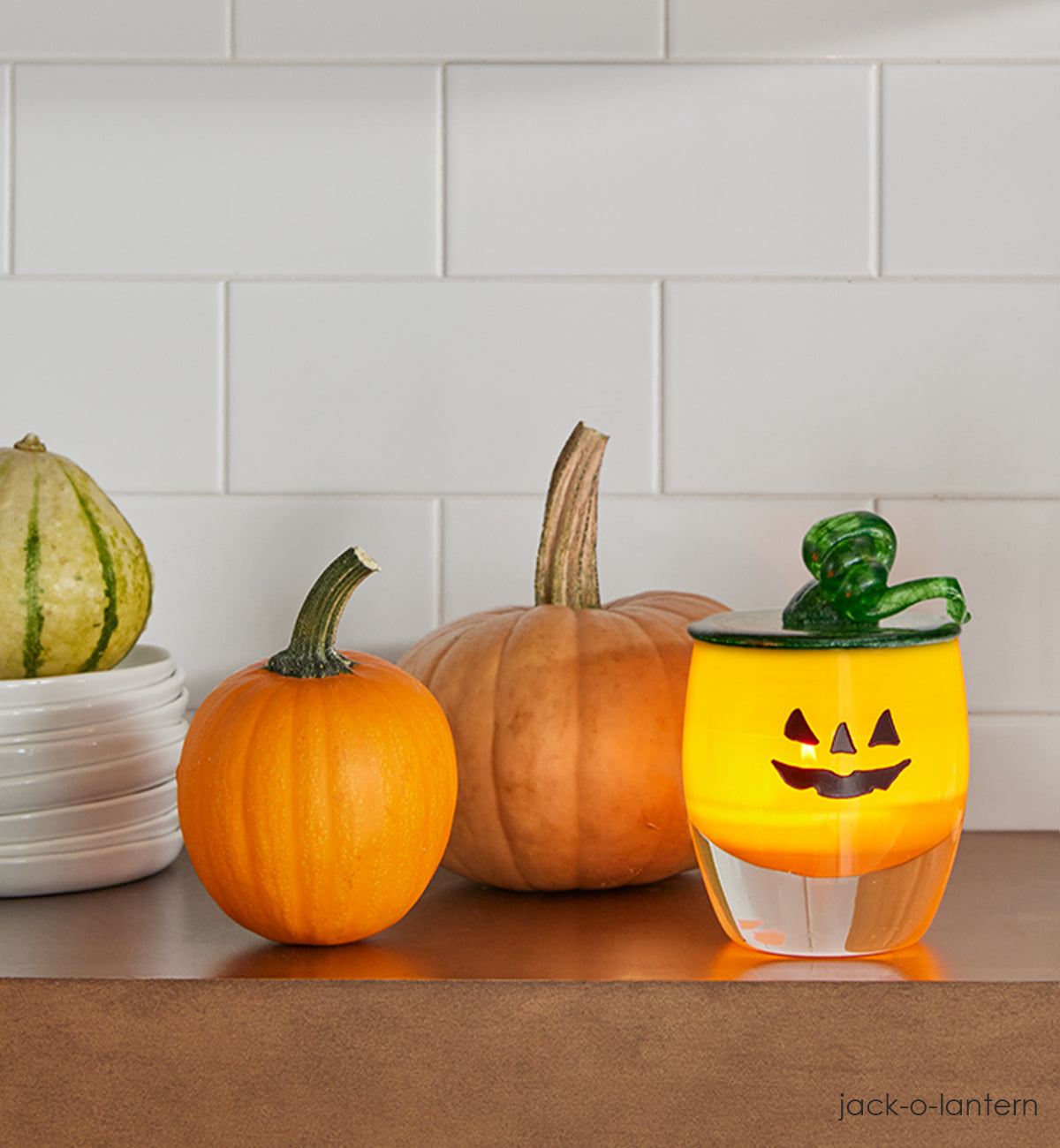 green seasonal snuffer, green hand-blown glass snuffer in pumpkin top and stem shape on a jack-o-lantern glassybaby on a kitchen shelf with gords. 