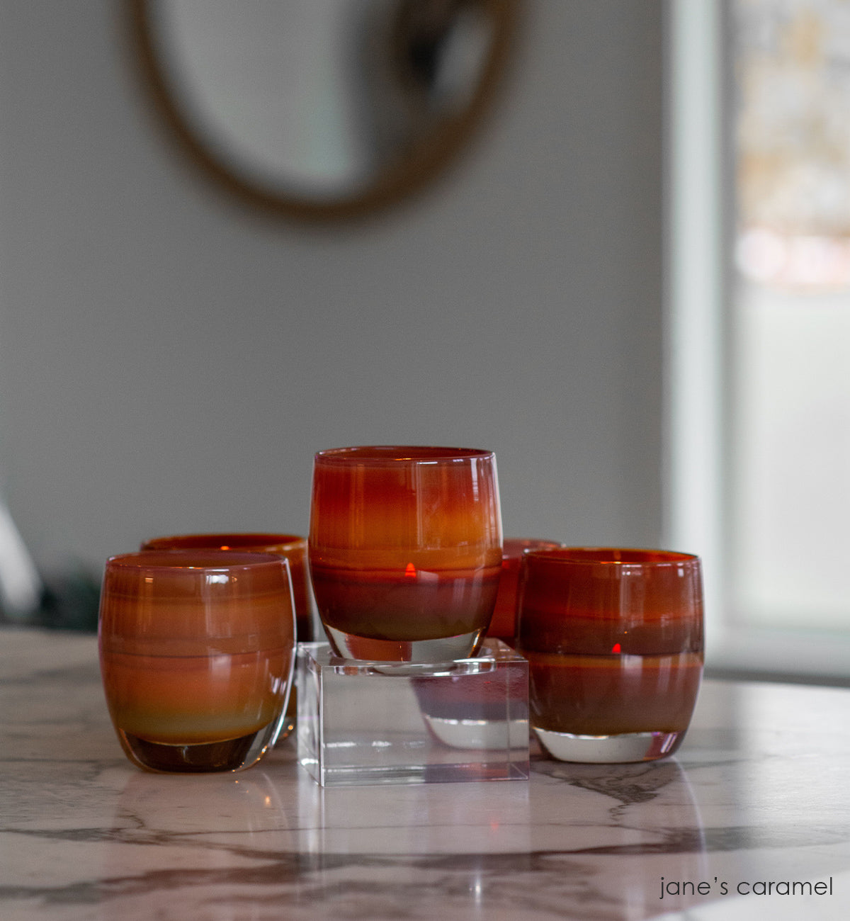 several jane's caramel, caramel brown and purple gradient, hand-blown glass votive candle holders, one on a baby stand on a black and white marble kitchen countertop with a mirror on the background wall.