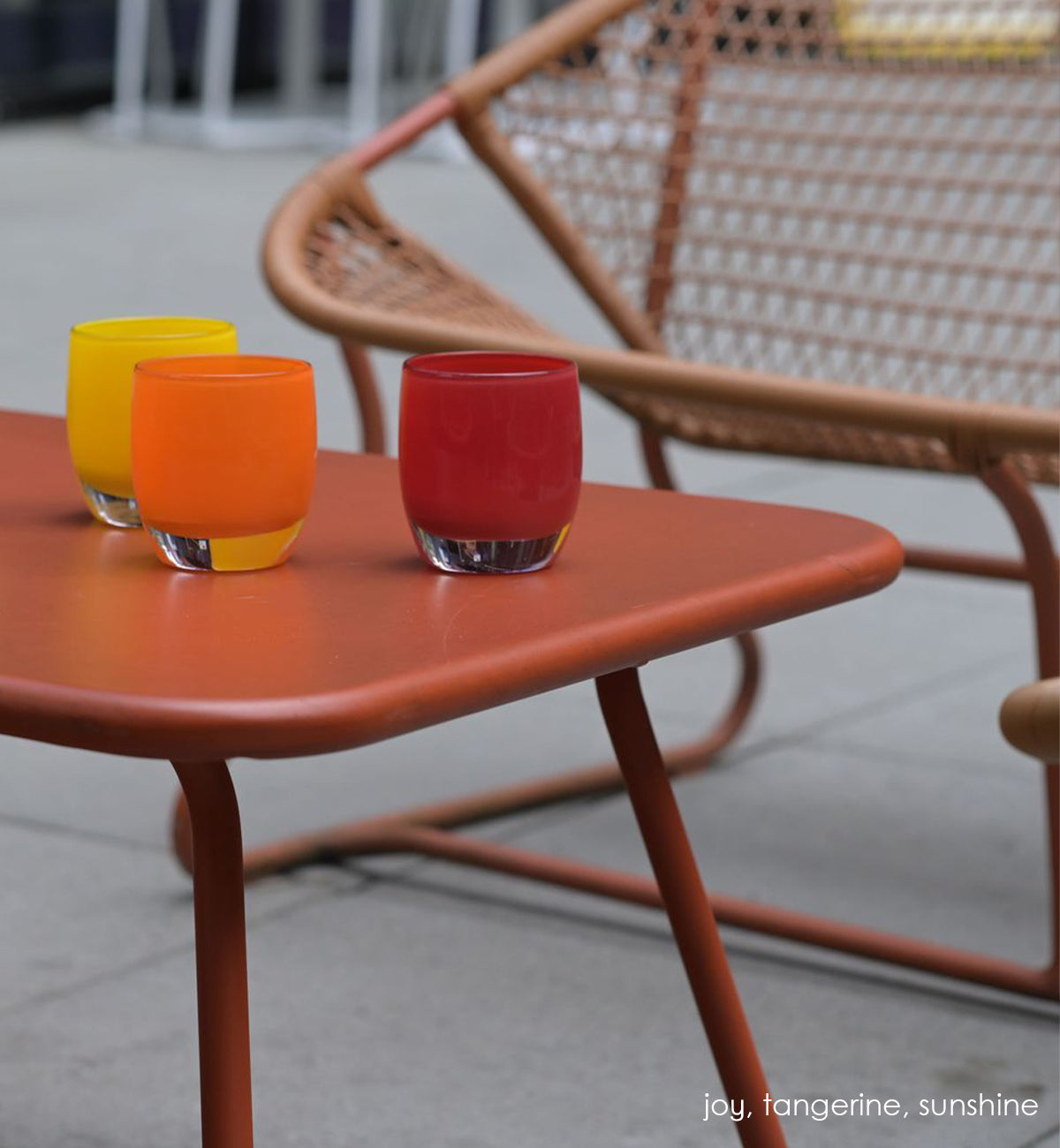 joy, crimson red hand-blown glass votive candle holder on a rusty orange outdoor patio table with tangerine and sunshine, with a netted bench blurred in the background.