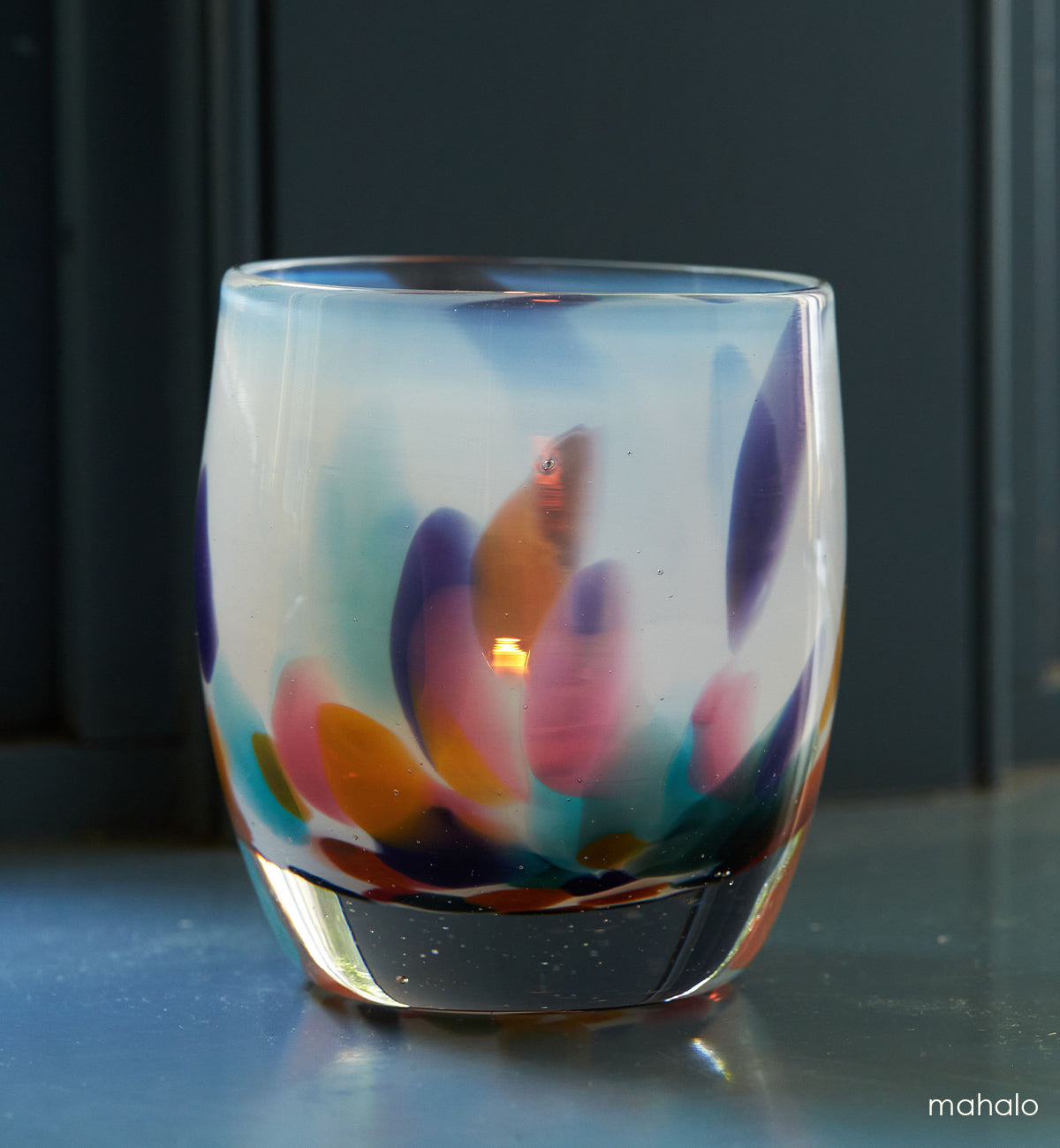 mahalo, white with multi-colored yellow, pink, teal, purple and green petals hand-blown glass votive candle holder on a blue grey surface.