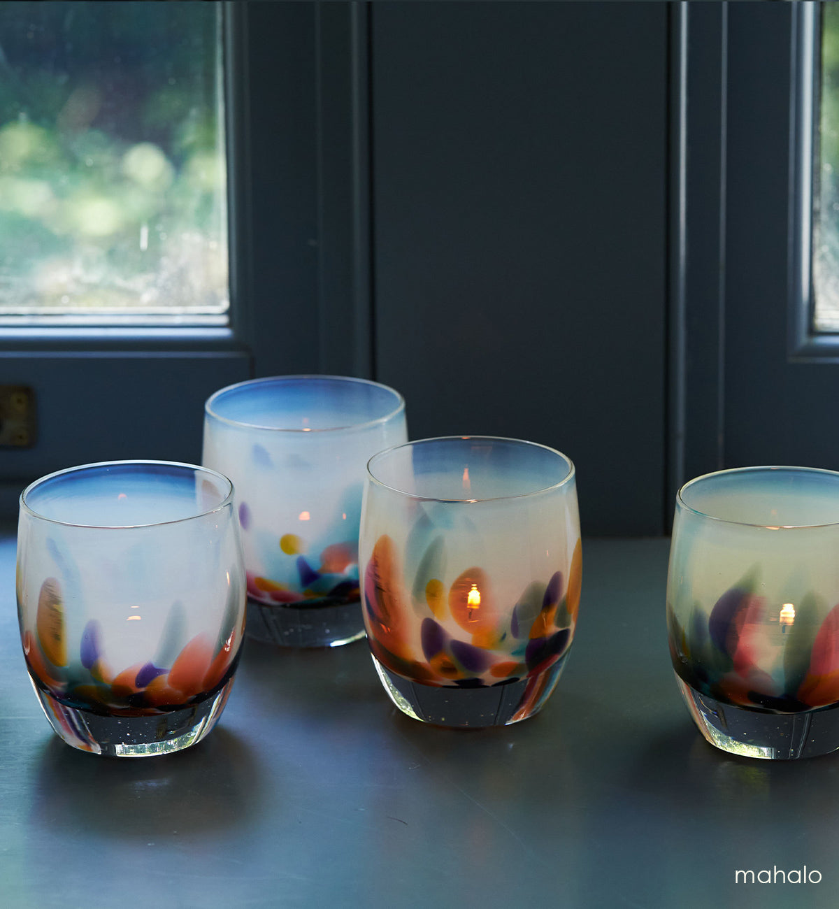 four mahalo, white with multi-colored yellow, pink, teal, purple and green petals hand-blown glass votive candle holders lit with tealights on a blue grey windowsill.