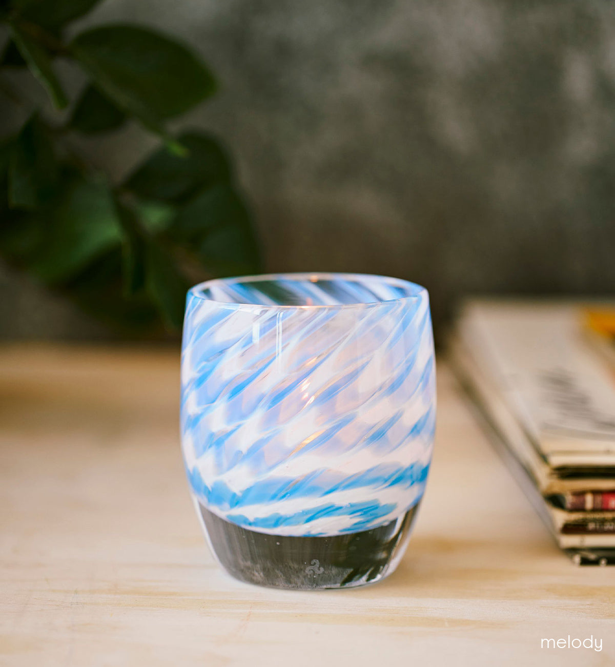 melody, sweeping swirled blue and white hand-blown glass votive candle holder on a light wood table with plant and stack of magazines in background.