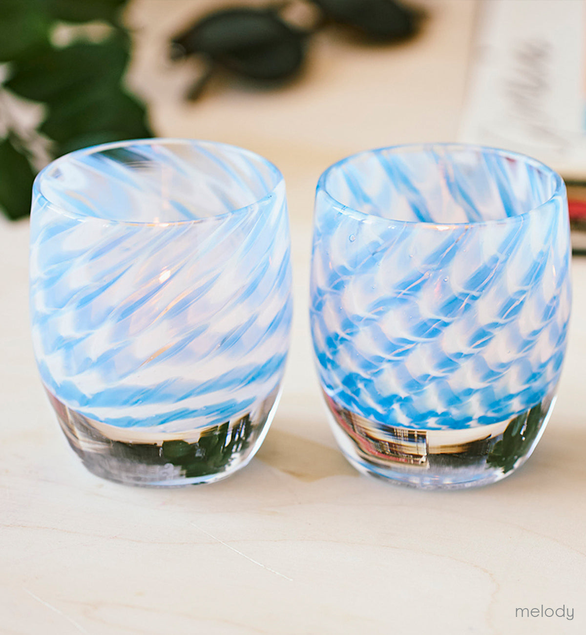two melody, sweeping swirled blue and white hand-blown glass votive candle holders on a light wood table with green plant and sunglasses in background.