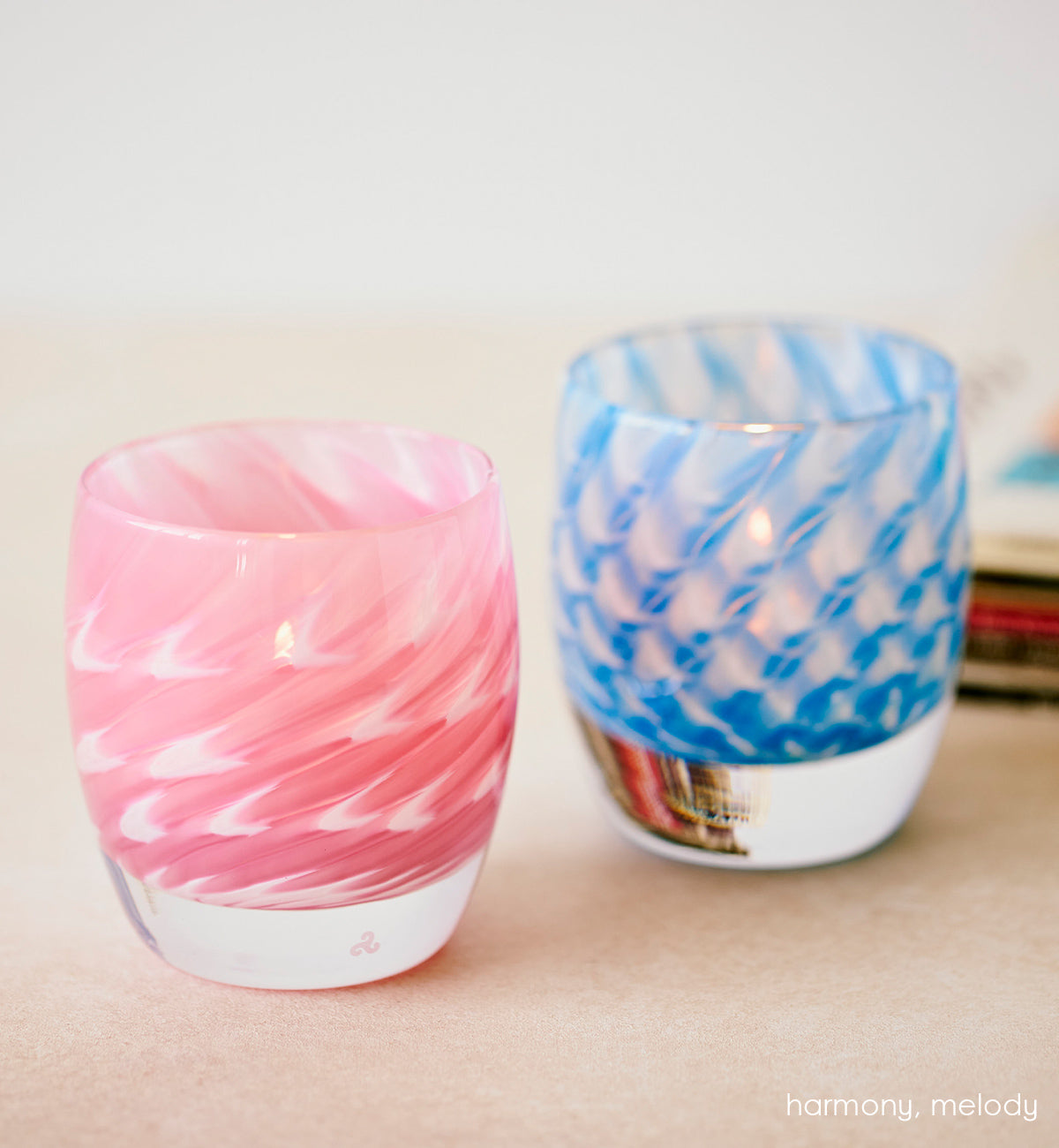 harmony and melody, sweeping pink and white and sweeping blue and white hand-blow glass votive candle holders on a table with stack of magazines behind.
