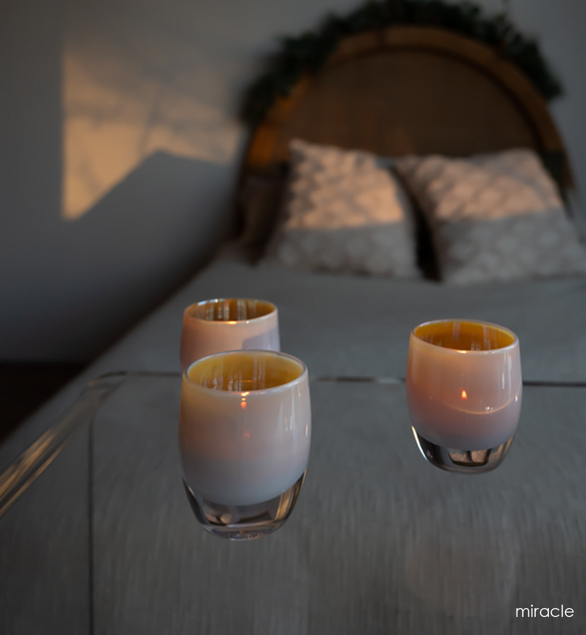 three miracle, golden beige double overlay metallic luster hand-blown glass votive candle holders on a clear acrylic table in front of a bed at sunset.