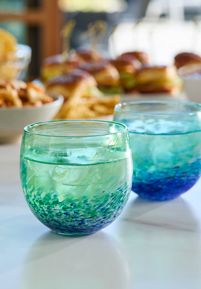 two SUNDAY drinkers, blue green and white swirl hand-blown drinking glasses with liquid and ice inside on a marble counter with a watch party spread of food in the background.