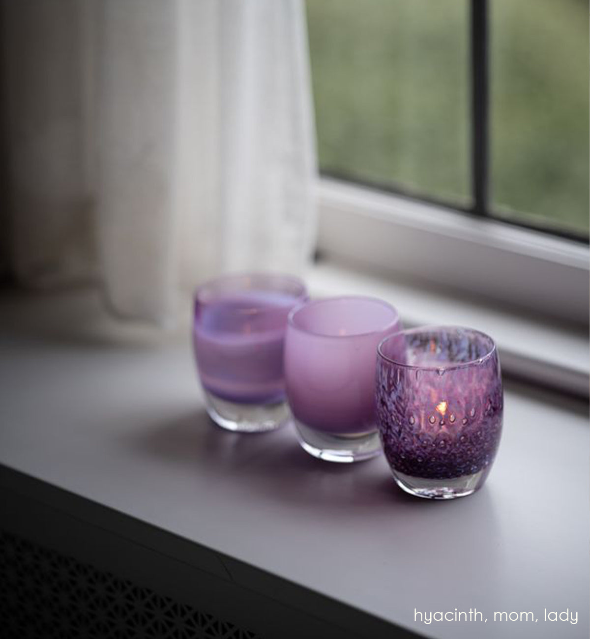 mom, soft purple hand-blown glass votive candle holder with hyacinth and lady sitting a gray windowsill next to a cream curtain.