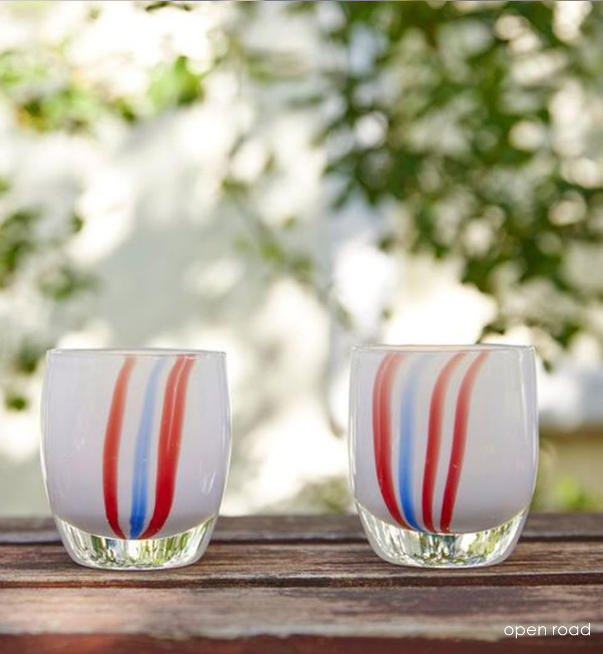 two open road, white with red and blue vertical striped hand-blown glass votive candle holders on a wood surface with sunshine and leaves in background.