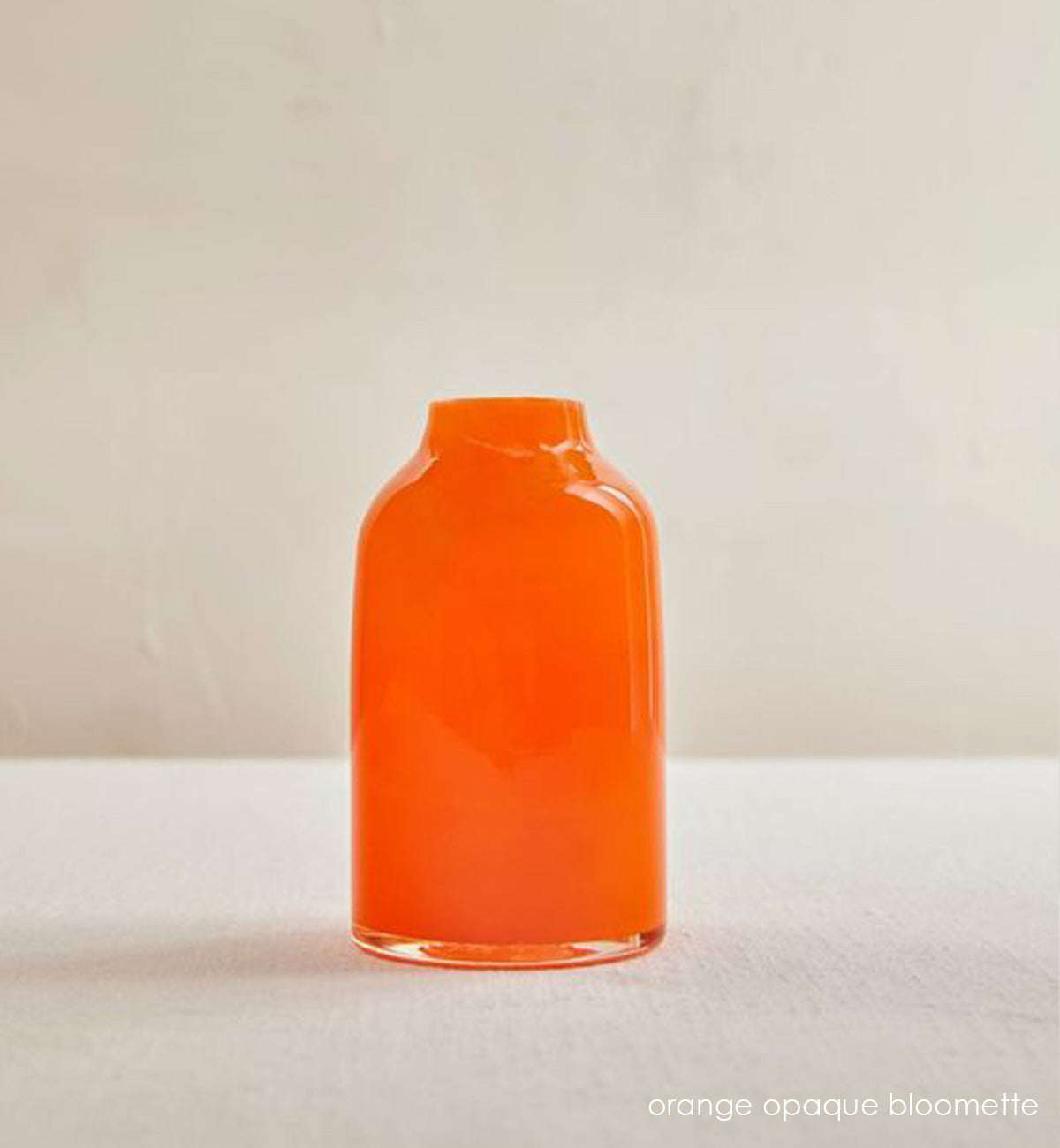 orange opaque bloomette, opaque orange hand-blown glass flower bud vase on cream table in front of cream wall.