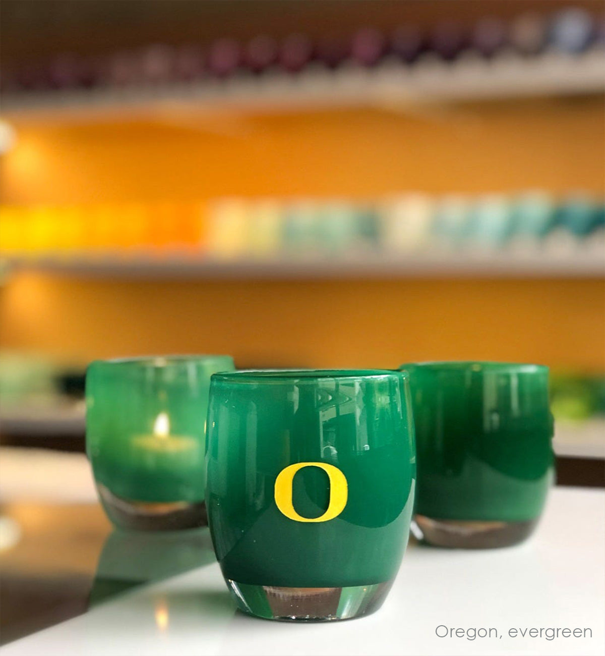 Oregon, green with sandblasted University of Oregon etching hand painted in yellow, hand-blown glass votive candle holder. Paired with evergreen on white and clear store table.