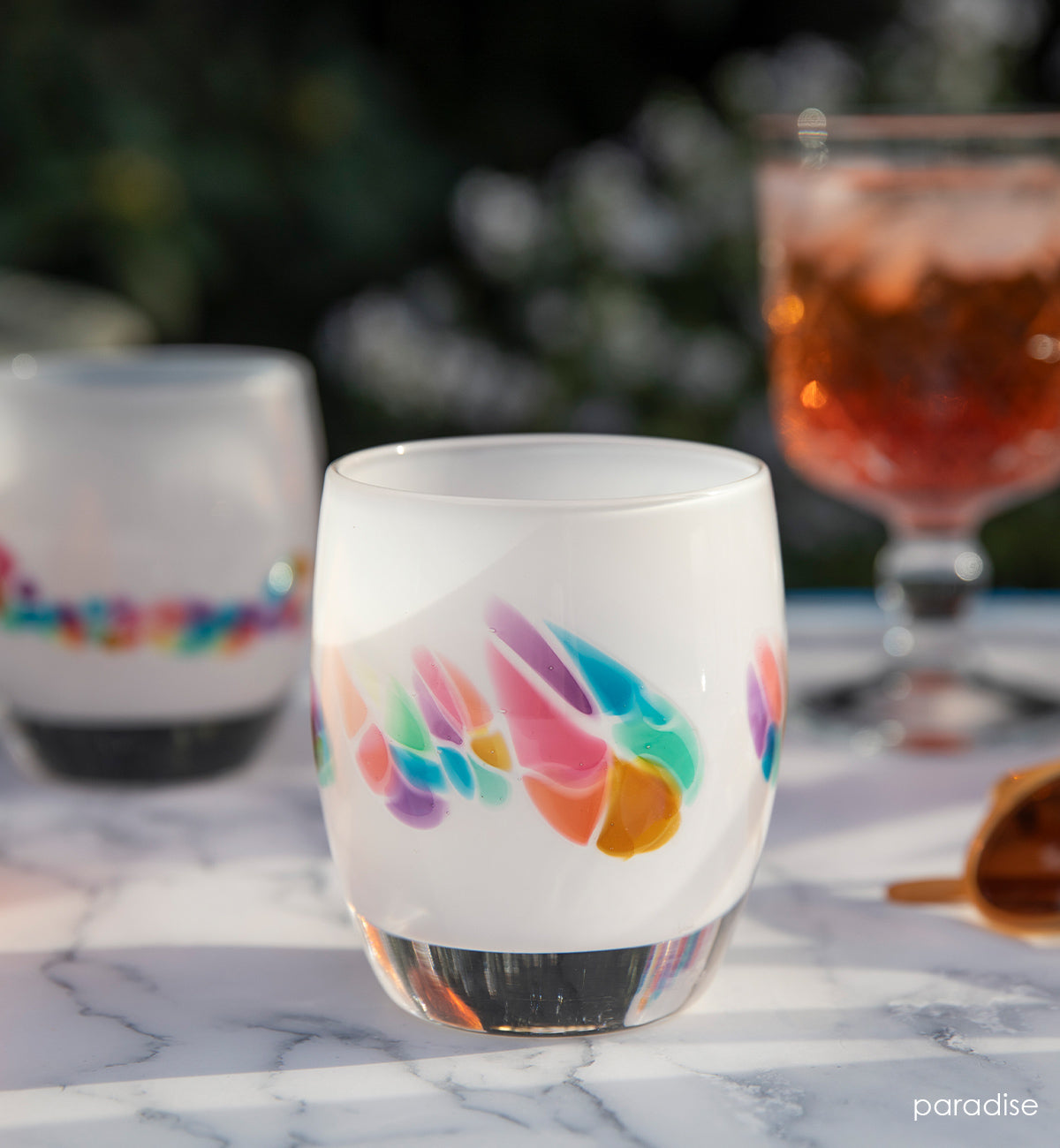 two paradise white rainbow murrini detail hand-blown glass votive candle holders on a marble table outside  with sunglasses and a wine glass.