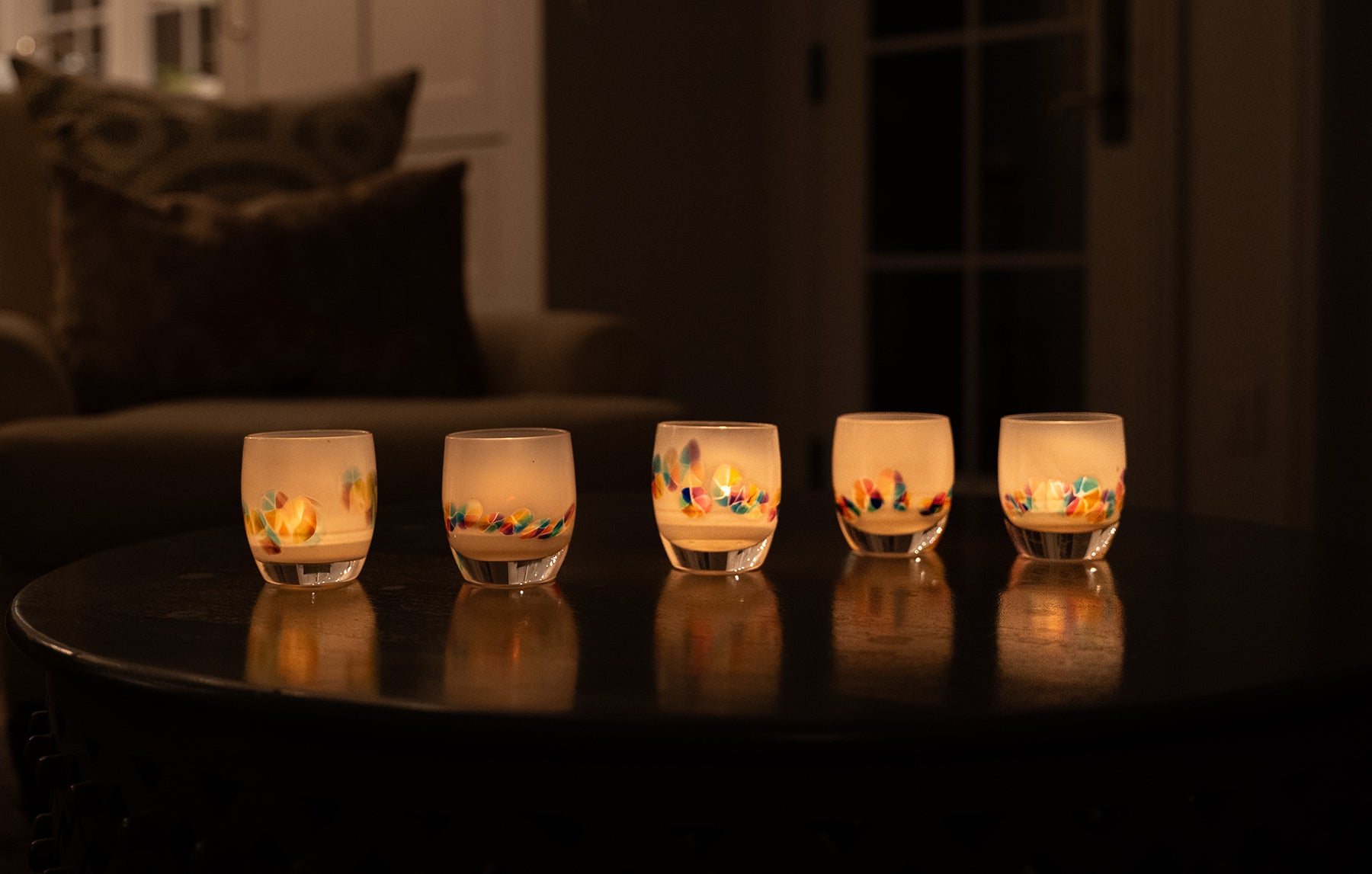 five paradise, white rainbow murrini detail hand-blown glass votive candle holders on a dark table, all lit and glowing with tealights.