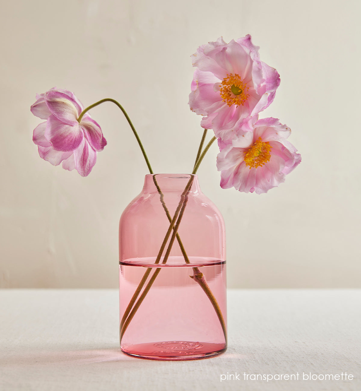 pink transparent bloomette, pink transparent hand-blown glass flower bud vase with pink and yellow flowers inside, sitting on cream table in front of cream wall.