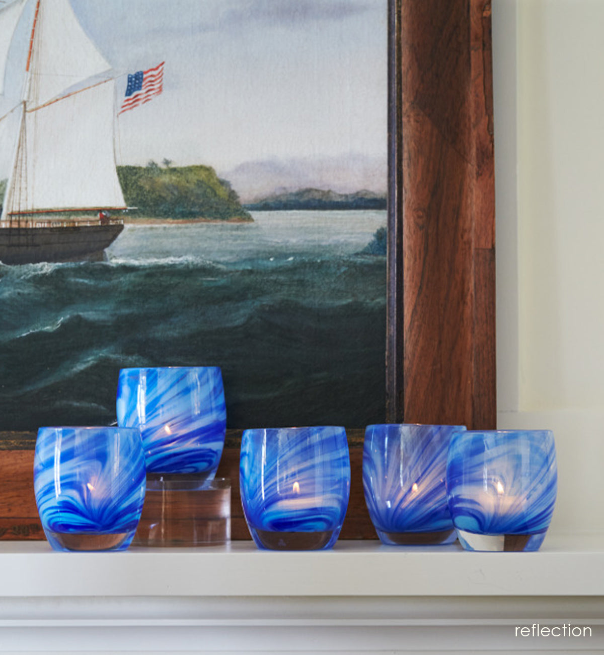 five reflection, transparent clear and blue raked hand-blown glass votive candle holders on a cream mantle with a painting of a ship behind.
