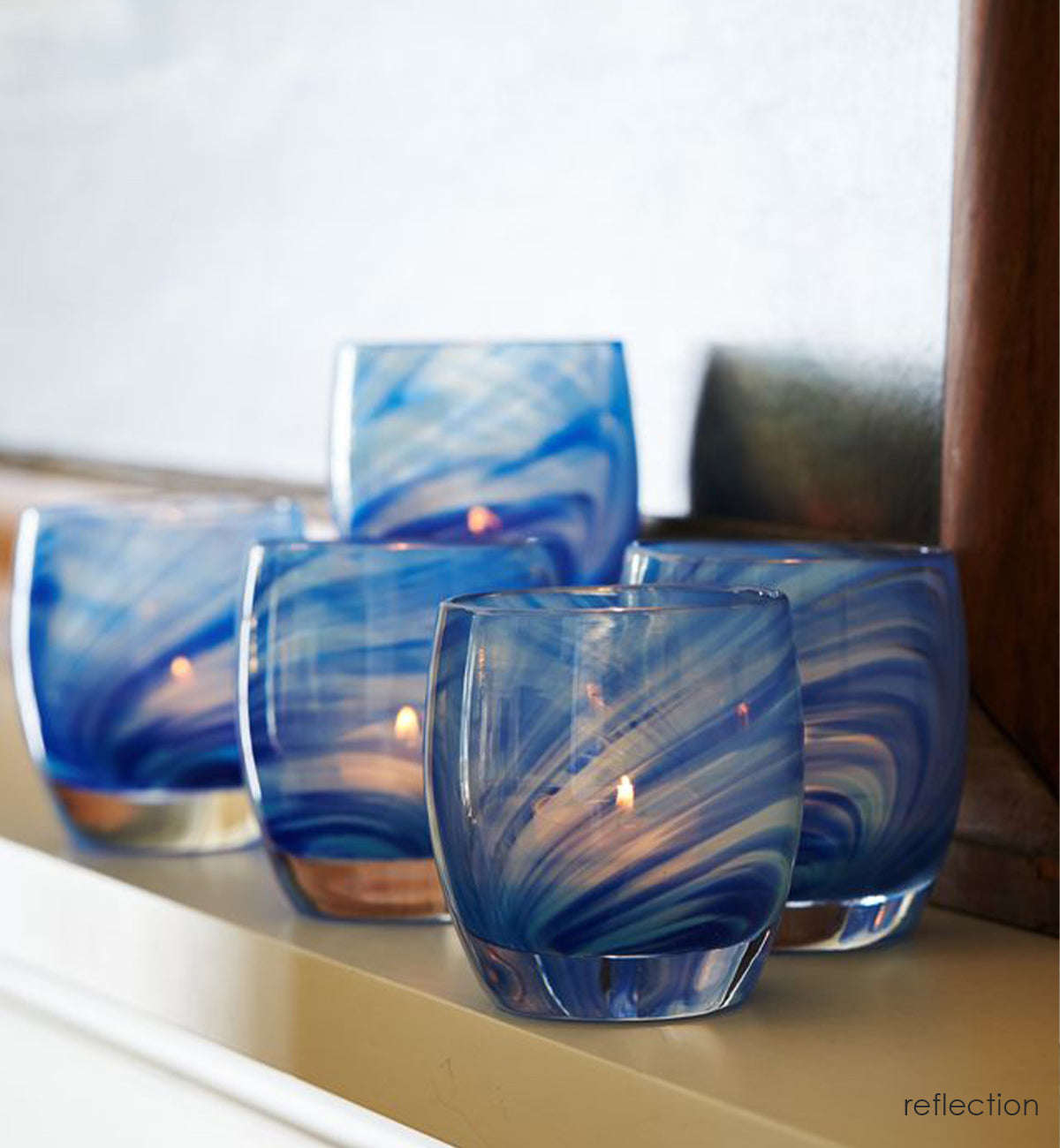 grouping of reflection, transparent clear and blue raked hand-blown glass votive candle holders on a mantle with lit tealights and daylight shining on them.