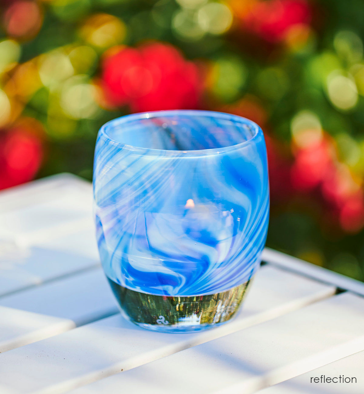 reflection, transparent clear and blue raked hand-blown glass votive candle holder on a patio table with red and green plants blurred in background.