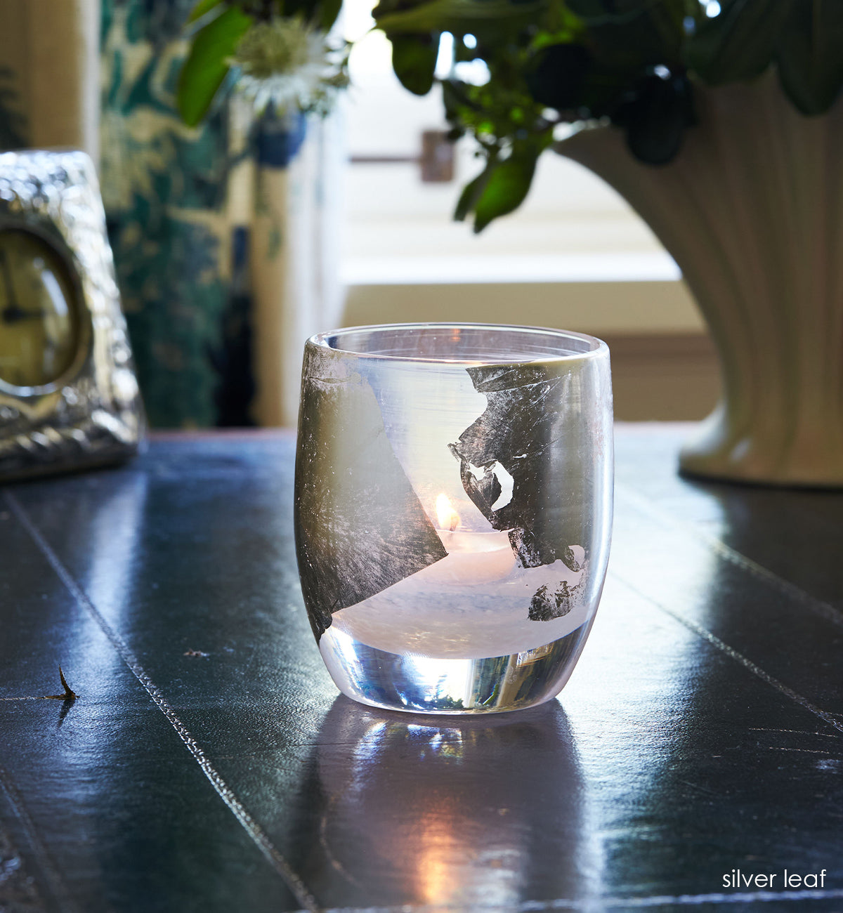 silver leaf, hand-blown glass votive cand holder with silver leaf with a lit tealight glowing inside. sitting on a navy blue desk with a decorative clock and vase in background.