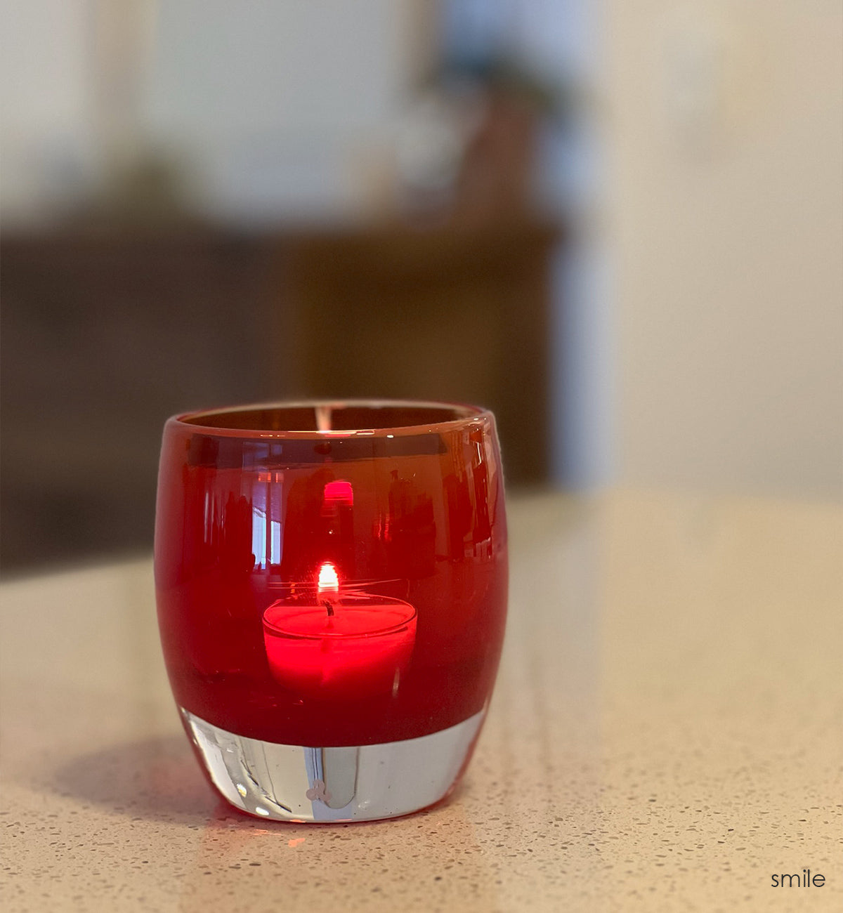 smile translucent bold red hand-blown glass votive candle holder. on a granite kitchen counter with wood furniture blurred in background.