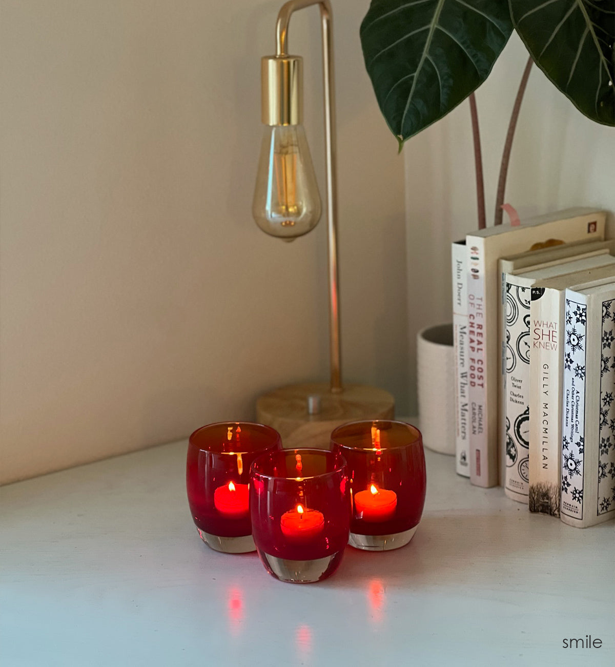 three smile, translucent bold red hand-blown glass votive candle holders with lit tealights on a desk with a lamp, plant and books.