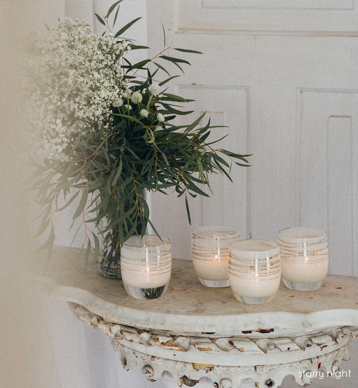 four starry night, white with silver shimmer wrap hand-blown glass votive candle holders sitting on a tall rustic table with  a vase of white flowers and green leaves in front of a white door.