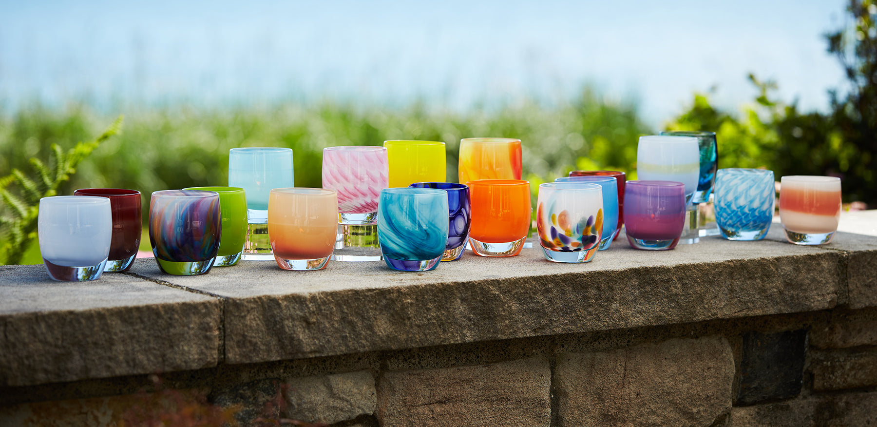 glassybaby summer collection, various summer glassybaby votives on a concrete stone wall with water and grass in background.