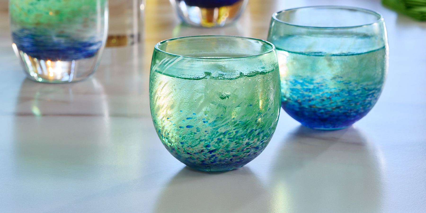 two SUNDAY drinkers, blue green and white swirl hand-blown drinking glasses with liquid and ice inside, on a white marble table with SUNDAY votives in background.