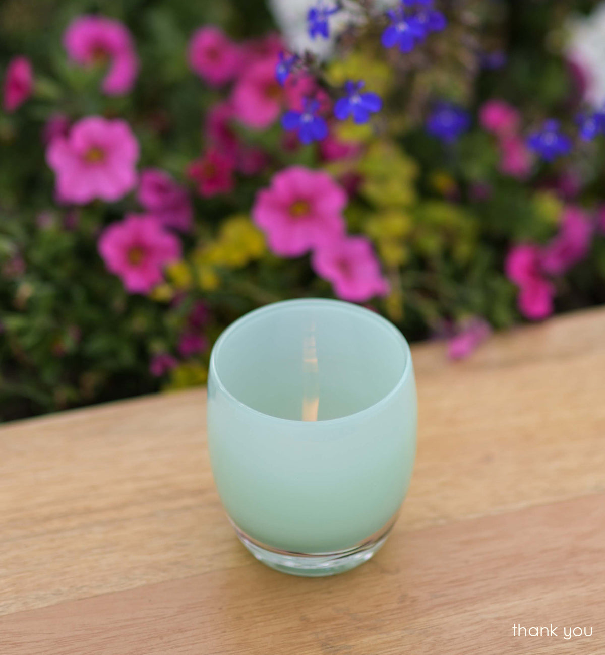 thank you mint green hand-blown glass votive candle holder on light wood table with pink and blue flower plants in background.