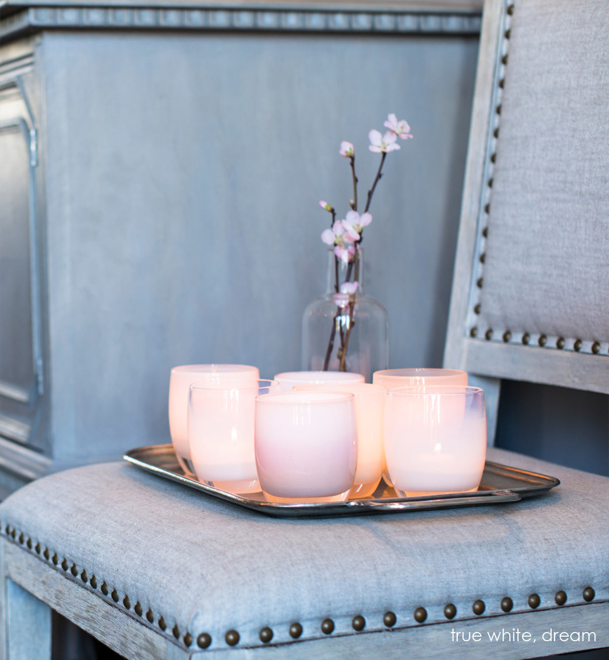 true white, opaque pinky white, hand-blown glass votive candle holder. Paired with dream on a metal chair sitting next to an orchid on a gray chair.