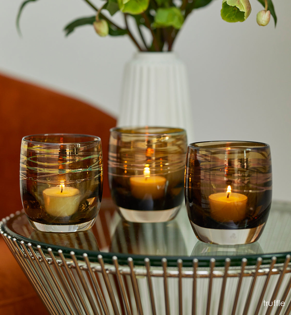 grouping of truffle, brown with metallic shimmer wrap hand-blown glass votive candle holders lit on a glass table. green plant in white vase in background.