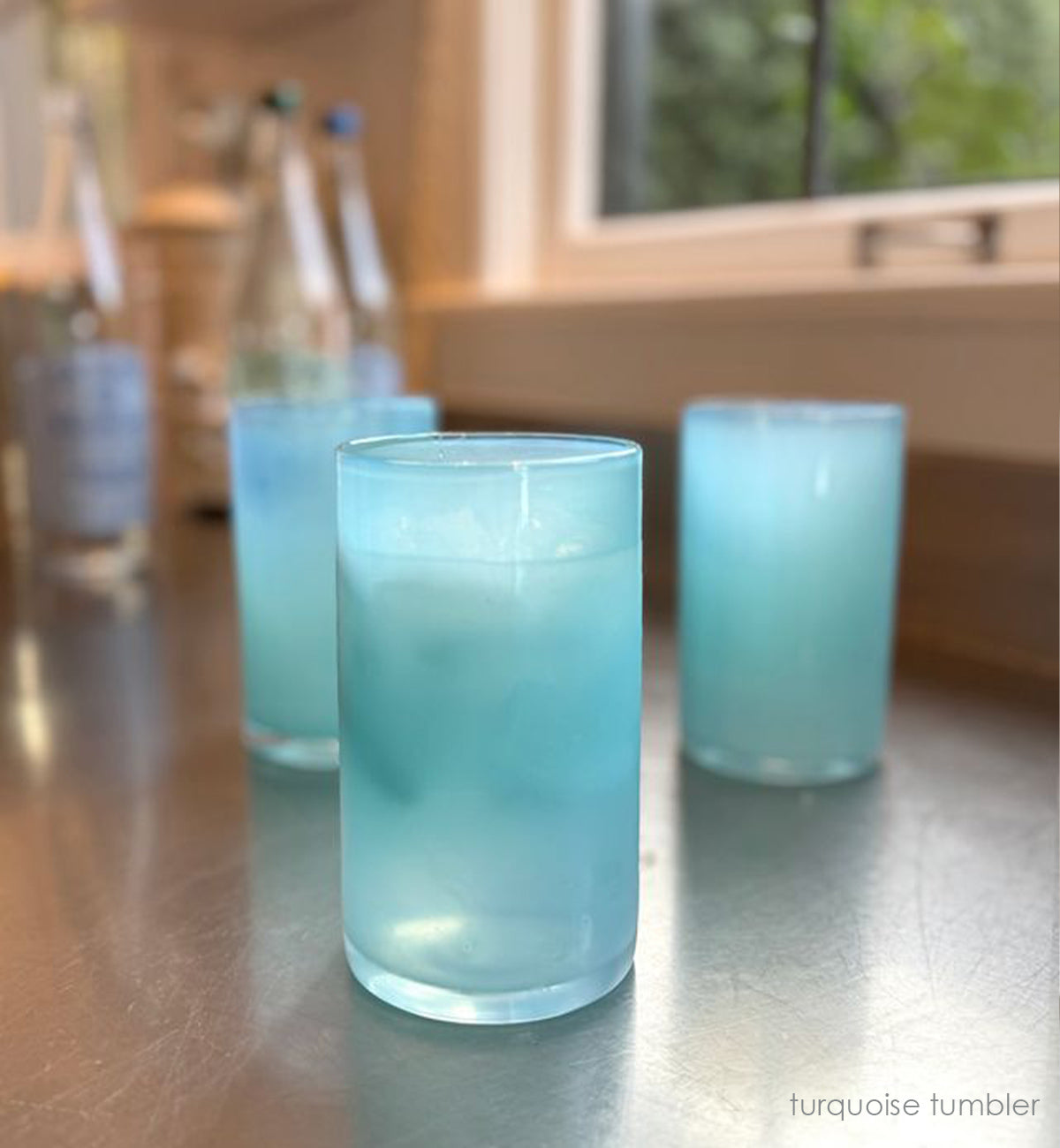 three turquoise tumblers, translucent ocean blue hand-blown water glasses on a counter with liquid and ice in each, and bottles in background.