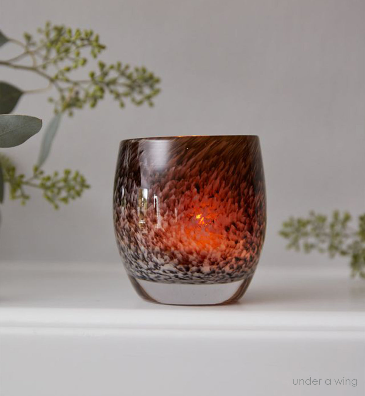 under a wing, an amber and brown mottled hand-blown glass votive candle holder on a white mantle with green plants on either side.