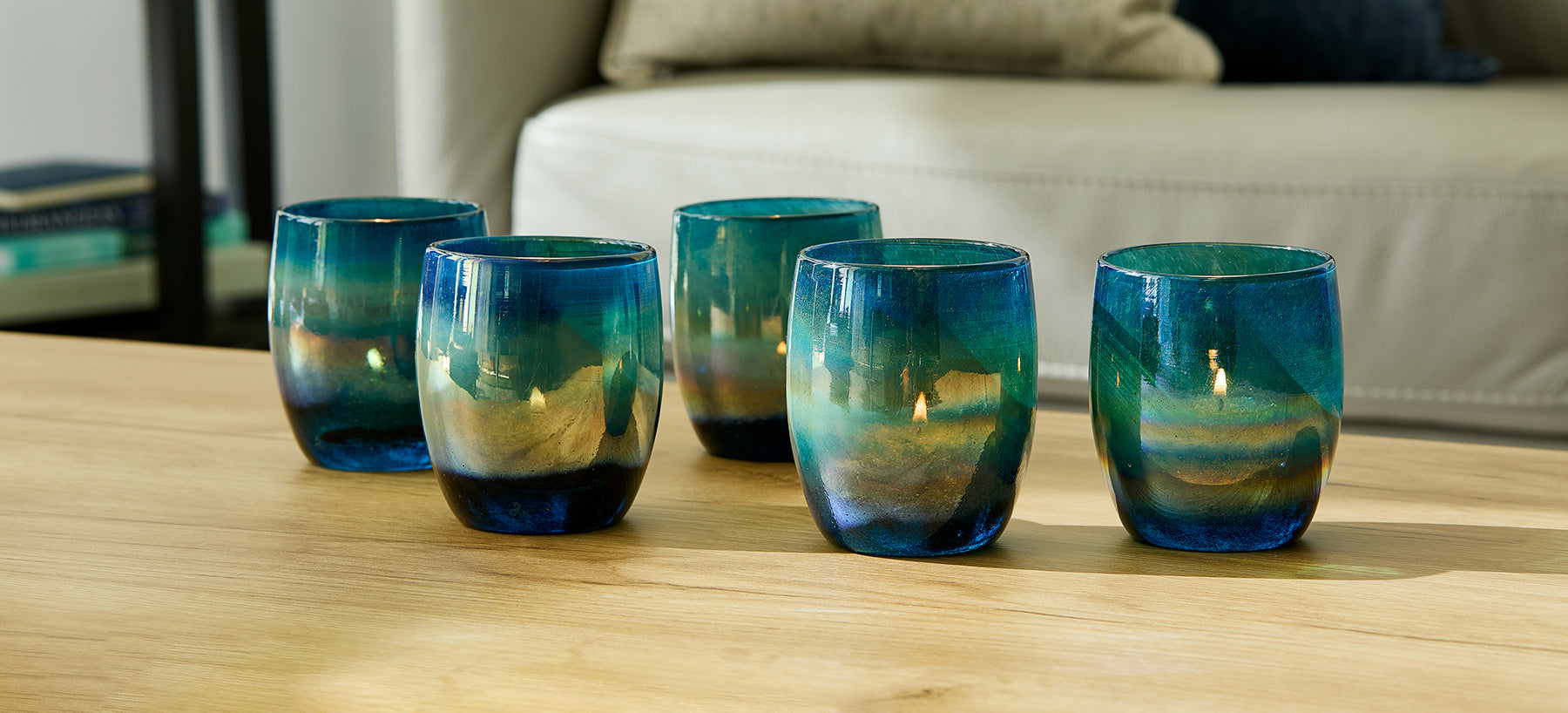 five under the sea, blue green metallic luster hand-blown glass votive candle holders lit with tealights on light wood table with gray couch in background.