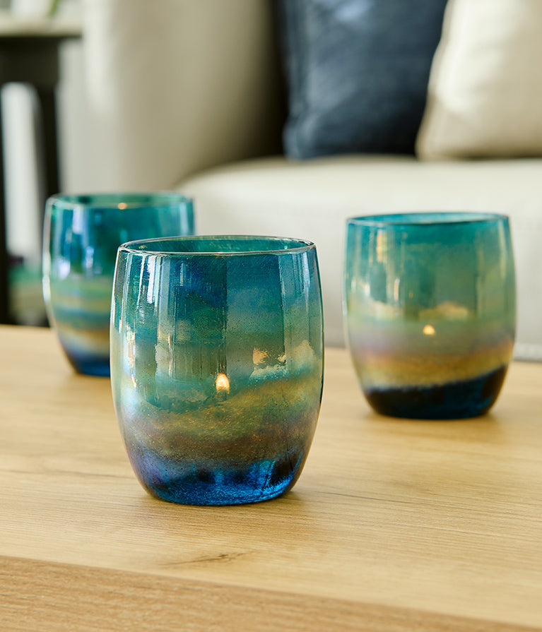 three under the sea, blue green metallic luster handblown glass votive candle holders on a light wood table with couch in background.