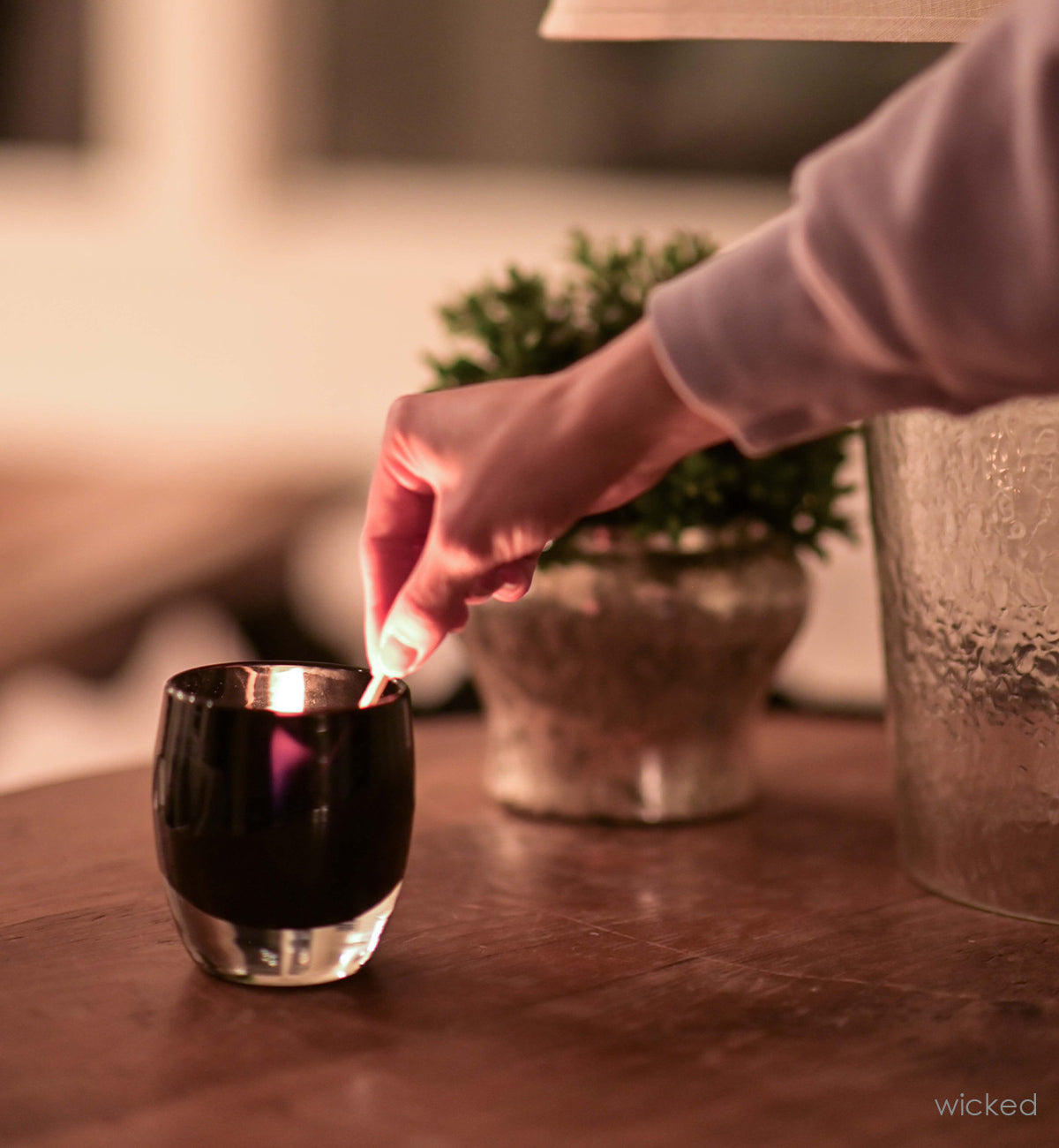 wicked, opaque purple black, hand-blown glass votive candle holder with a tealight being lit with a match inside, placed on a wood table with a lamp and potted plant. a purple flame glowing.